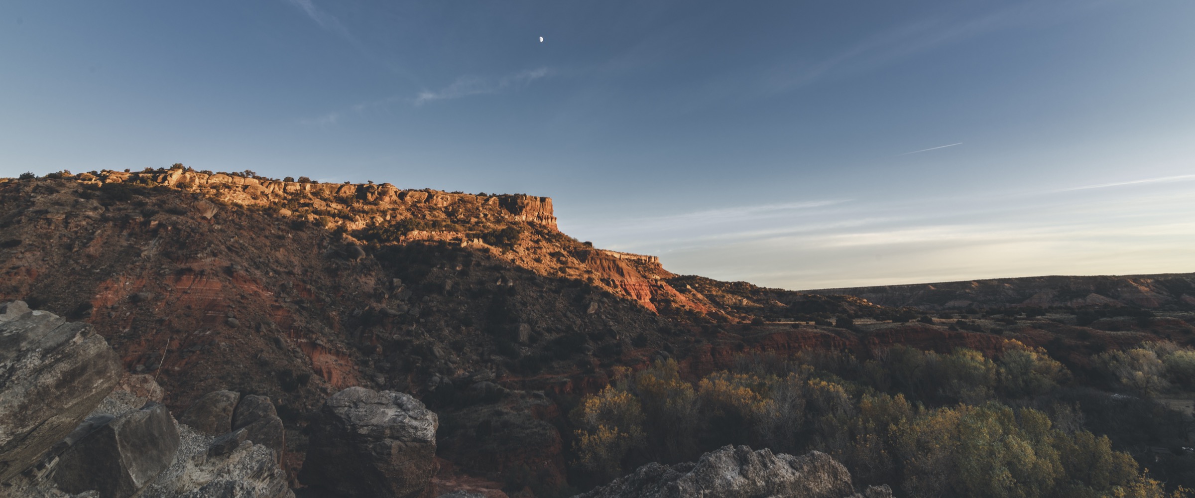 Panoramic view across a canyon valley, with sunlit cliffs and trees below a wide blue sky.