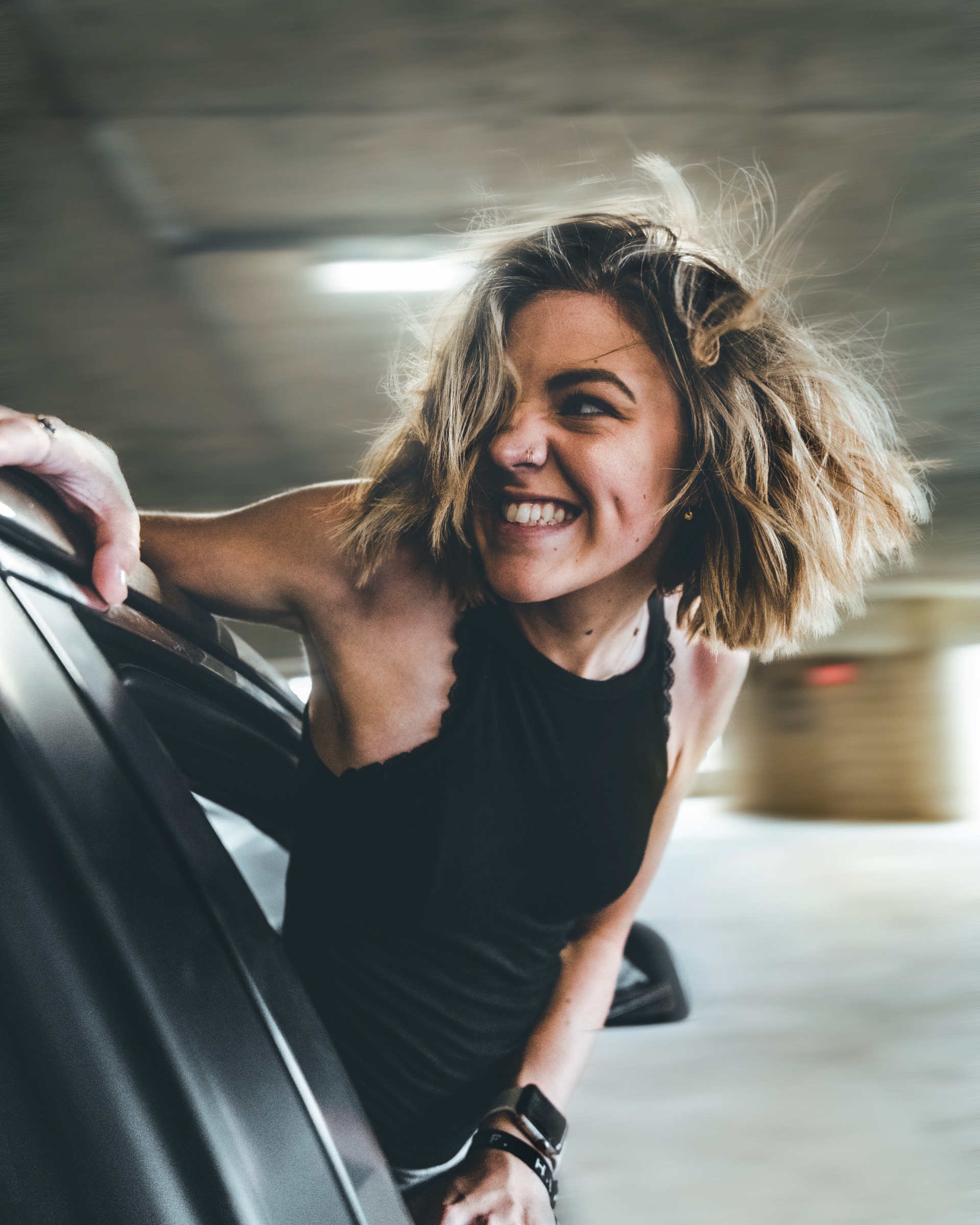 Smiling person leaning beside a car in a parking garage, wearing a sleeveless black top.